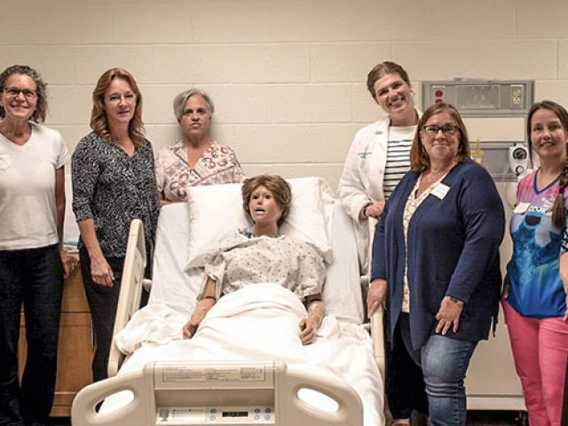 Seven IMPART Alliance Train the trainer students standing next to our assistant director and training instructor Bethany Duyser. They are standing, surrounding a manikin.