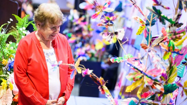 An older woman wearing a red cardigan smiles while looking at a colorful art installation made of paper butterflies and flowers arranged on branches in an indoor exhibit space.