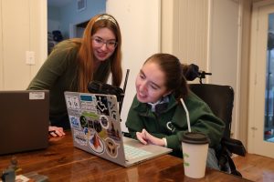 Two young women sit together at a computer, collaborating on a project. One woman with long brown hair and glasses leans over the other’s shoulder, while the second woman, wearing a green top, looks closely at the laptop screen. The laptop has several stickers on its cover, and a camera is positioned nearby.